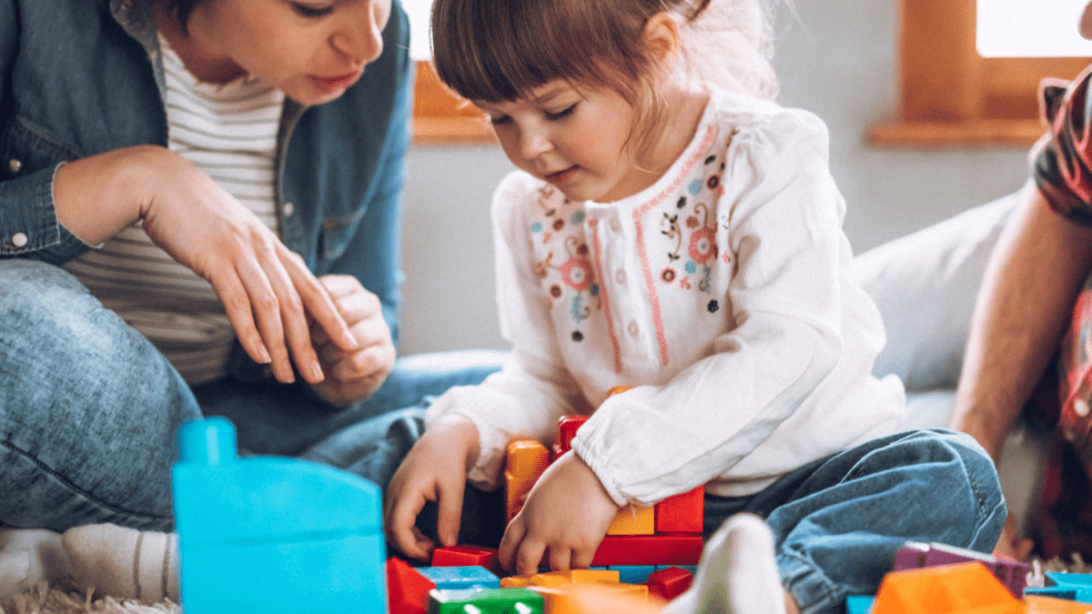 A woman (either a mother or caregiver) playing with a girl with building blocks.